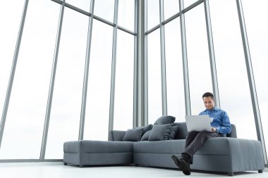 Genç Asyalı işadamı kanepede otururken dizüstü kullanarak. Mutlu gülümseme ve bir fincan el altında. Modern ofis büyük penceresinde