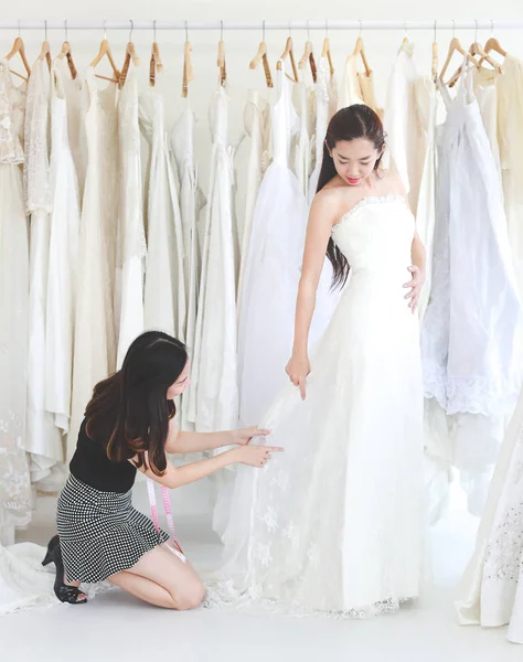 An young woman is trying wedding dress in studio, designer is assisting, nice standing posting, some dresses backgound.