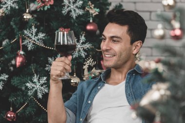 Genç yakışıklı kot gömlek arıyor beyaz kırmızı şarap cam modern oda için arka plan olarak dekore edilmiş bir Noel ağacı var.