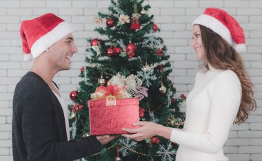 Genç ve yakışıklı beyaz adam ve güzel bir kadın ayakta birlikte tutarak büyük hediye kutusu önünde büyük bir Noel ağacı süslü. Sevgili Noel Festivali iyi zaman paylaşımı için kavramı.