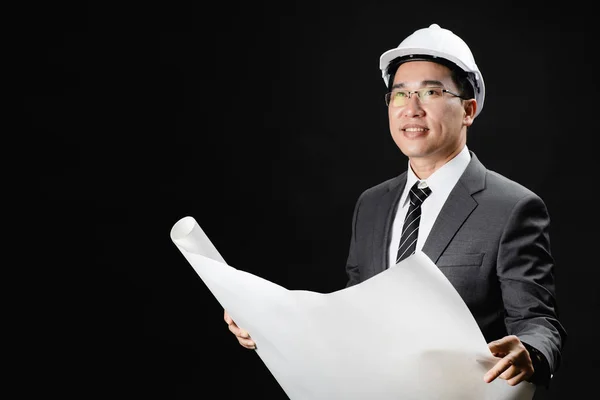 Portrait of Asian business man in suit and tie, wearing a white safety hat, looking at  construction plans in hand, confidence and trustworthy. Isolated in black backgound.