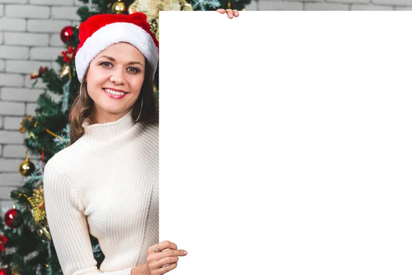 Beautiful and young caucasian woman holding and pointing to blank white board in front of big christmas tree. Concept for advertising  in christmas festival.