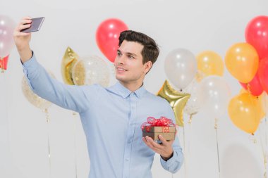 İyi bir hediye kutusu tutan genç beyaz adam eğlenceli mutlu selfie parti, beyaz arka plan ile şenlikli renkli balonlar.