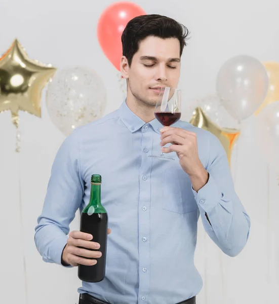 Attractive young caucasian man, tesing wed wine from glass feeling happy at work party, white background with festive colorful balloons.