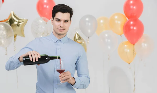 Handsome young caucasian man pouring red wine in to glass at work party, portrait against white background with colorful festive balloons.