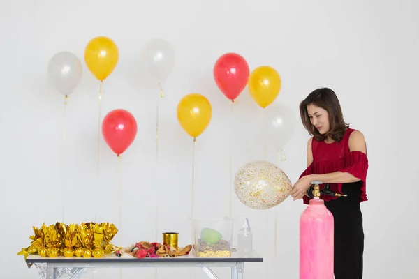 altın kağıt, dekorasyon ürünler line-up masada, hediyelerle kaydırma kırmızı bluz ve siyah pantolon helyum gaz tank ile renkli balon şişirme giyen genç güzel Asyalı kadın hazırlamak için parti,