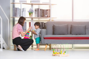 Mutlu Asya Aile, Genç, güzel annesi pembe bluz ve vakit, çok renkli ahşap masa oyuncakları rahat oturma odasında oynarken Gri tişört, yeşil kısa, sevimli küçük yürümeye başlayan çocuk