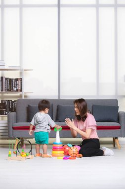 Mutlu Asya Aile, Genç, güzel annesi pembe bluz, yere, oturma içinde sarı ring, gri T-shirt, kısa, sevimli küçük yürümeye başlayan çocuk zevk topla oynamayı ve oyuncaklar oturma odası rahat tutun