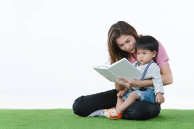 Asyalı bir aile, evde mutlu vakit geçirme zamanı, yapay yeşil çimlerde oturan genç bir anne, şefkatle kucağında sevimli, tapılası bir bebek, onun için beyaz arka planda kitap okuyor.