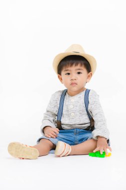Stüdyo portresi, şirin, sevimli, Asyalı, kot pantolon giyen, hasır şapkalı, uzun kollu tişörtlü, yerde oturan, oyuncak oynayan, izole beyaz arka plandaki kameraya bakın.