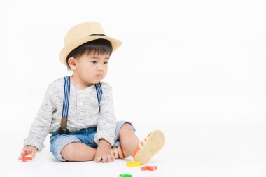 Stüdyo portresi, şirin, sevimli, Asyalı, kot pantolon giyen, hasır şapkalı, uzun kollu tişörtlü, yerde oturan, oyuncak oynayan, izole beyaz arka plandaki kameraya bakın.