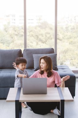 Asian family, happy time at home, young beautiful mother, sitting on floor, cute, adorable toddler, on her laps, affectionately pointing something on notebook screen for him, with sofa background clipart