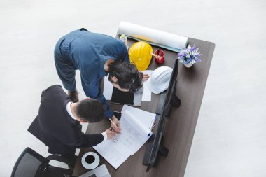 Siyah takım elbiseli Asyalı iş adamının en iyi görüntüsü, çizim yapmakla meşgul, ustabaşıyla konuşuyor, kırtasiye malzemesi, beyaz ve sarı güvenlik kaskı, dizüstü bilgisayar, tablet, akıllı telefon, ofis geçmişi.