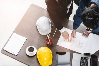 Yakın plan, Asyalı iş adamı ve erkek ustabaşının üst görüntüsü, çizim, bina planı tasarımı, dizüstü bilgisayar, miğfer ve çalışma masasında tartışmalar..