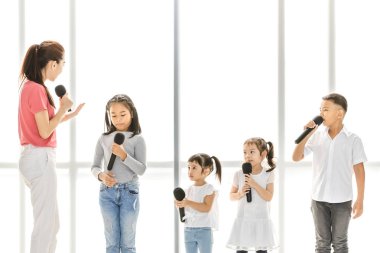 Asyalı öğretmen öğretmek bir şarkı söylemek Asyalı çocuklar, çocuklar onu dinlemek dikkat, büyük beyaz pencere önünde duruyorlar. Müzik okulu veya şarkı sınıf eğitimi kavramı.