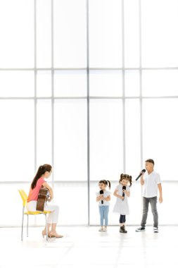 Asyalı öğretmen akustik gitar bir şarkı söylemek Asyalı çocuklar öğretmek bir sandalye oyunundan oturan çocuklar onu dinlemek dikkat, onlar büyük beyaz pencere önünde stand.