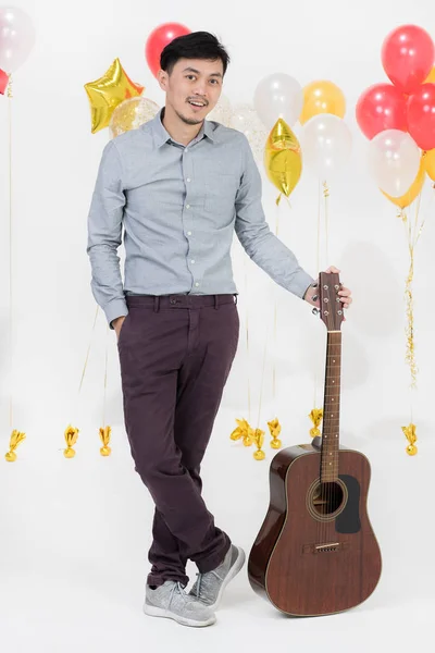 Full-length studio portrait of Asian young man, black hair, in gray shirt, smiling,look straight to camera,holding acoustic guitar on party scene with bunch of balloons on isolated white background