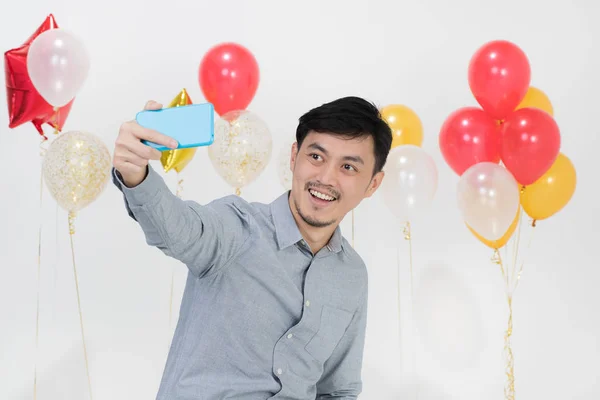 Portrait of handsome Asian young man, black hair, in gray shirt, joyfully smiling, taking selfie with smartphone  on party scene with bunches of balloons on isolated white background