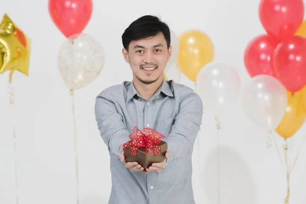 Half-length studio portrait of Asian young man,black hair,in gray shirt,smiling,hold out hands with small present box,on party scene on isolated white background