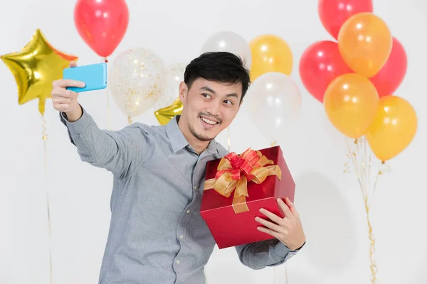 Portrait of Asian young man, black hair, in gray shirt,joyfully smiling, holding red present box,taking selfie with smartphone on party scene with bunches of balloons on isolated white background