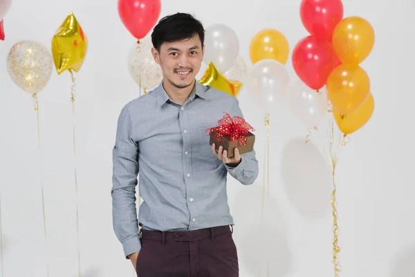 Half-length studio portrait of Asian young man,black hair,in gray shirt,smiling,right hand in pant pocket , left hand holding small present box, on party scene on isolated white background