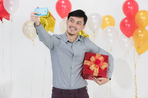 Portrait of Asian young man, black hair, in gray shirt,smiling, holding red present box under arm,taking selfie with smartphone on party scene with bunches of balloons on isolated white background