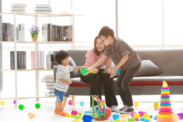 Asian family, happy time at home, young beautiful mother, taking care sons, little adorable toddler, standing, young boy wearing eyes glasses hold green ball, playing together in modern living room