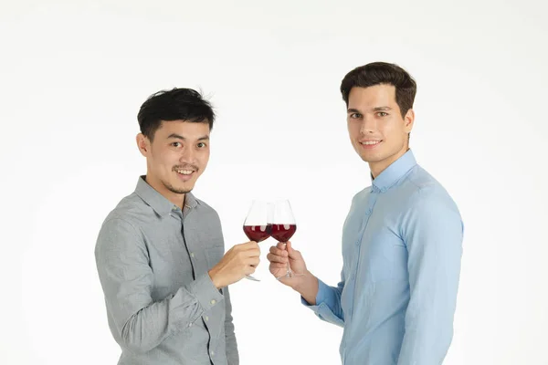 Half-length studio portrait of two young, handsome men, smiling and looking at camera, clinking wine glasses in celebrating some special occasion, standing on isolated white background