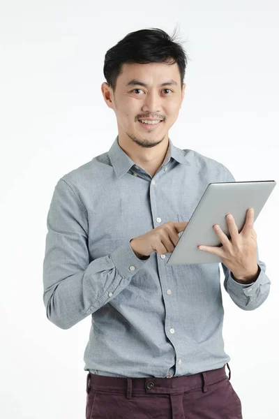 灰色シャツ 保持および分離の白い背景の上のカメラを見て 顔に笑みを浮かべて デジタル デバイス上で何かを指しての若いアジア男のハーフレングス スタジオ ポートレート — ストック写真