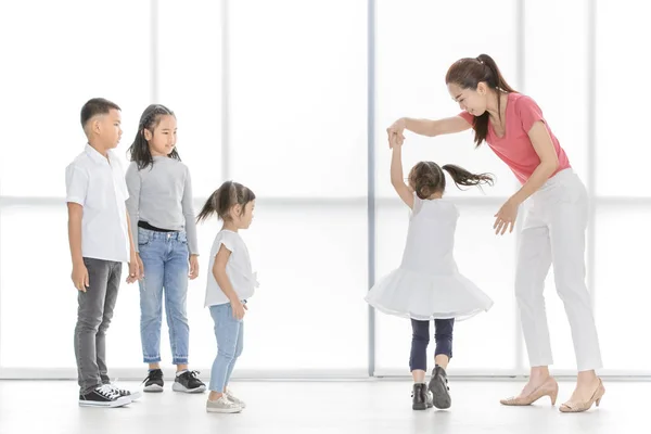 Asian woman in pink shirt dance with little Asian girl, Asian boy and gilrs look at them, they stand in front of big white window.