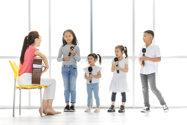 Asyalı öğretmen akustik gitar bir şarkı söylemek Asyalı çocuklar öğretmek bir sandalye oyunundan oturan çocuklar onu dinlemek dikkat, onlar büyük beyaz pencere önünde stand.