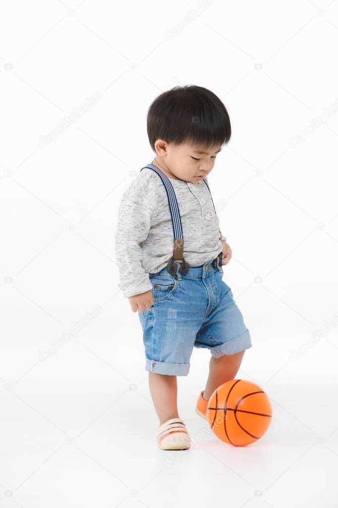 Studio portrait of adorable, Asian toddler boy wearing denim overalls, long sleeve T-shirt, orange shoes, standing, look down and playing with orange plastic ball, on isolated white background