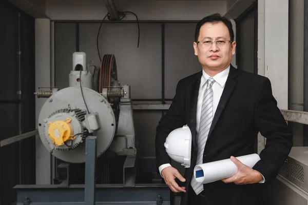 Asian executive engineer stand beside heavy machinery in industrial factory, he holds white helmet in his arm,concept for industrial factory.