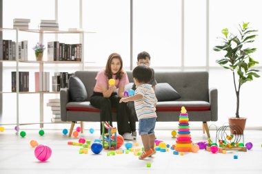 Asyalı aile, evde mutlu zaman, genç güzel anne, oğulları ile oynarken, küçük sevimli yürümeye başlayan, modern oturma odasında gözleri gözlük takan genç çocuk etrafında dağınık çok renkli oyuncaklar dolu
