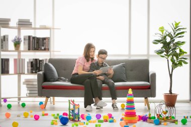 Asya Aile, mutlu, boş zaman zaman evde, genç güzel anne ve genç oğlu koltukta oturan, kitap okumak, ödev modern oturma odası renkli oyuncaklar tam olarak öğretim gözler gözlük