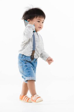 Stüdyo portresi, şirin, tapılası Asyalı, kot pantolon giyen, uzun kollu tişört giyen, turuncu ayakkabı giyen, yürüyen, konuşan ve akıllı telefonuyla beyaz arka planda oynayan çocuk.