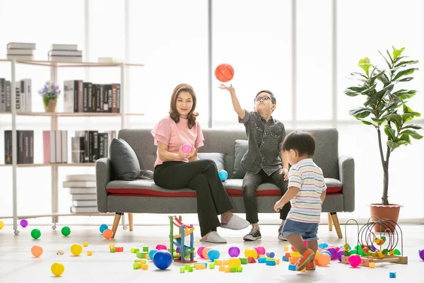 Asian family, happy time at home, young beautiful mother and young son wearing eyes glasses sitting on sofa, throwing ball, cute little toddler walking around playing together in modern living room
