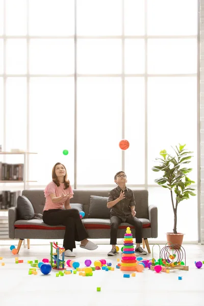 Asian family, happy time at home, young beautiful mother and young son wearing eyes glasses sitting on sofa, throwing ball, cute little toddler walking around playing together in modern living room