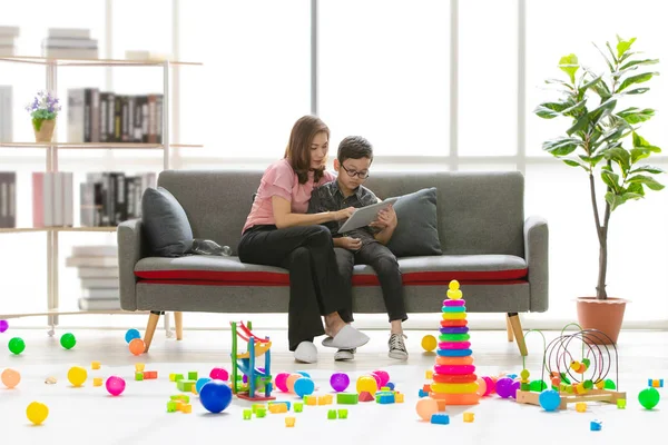 Asian family, happy, leisure time at home, young beautiful mother and young son wearing eyes glasses sitting on sofa, reading book, teaching homework in modern living room full of colorful toys