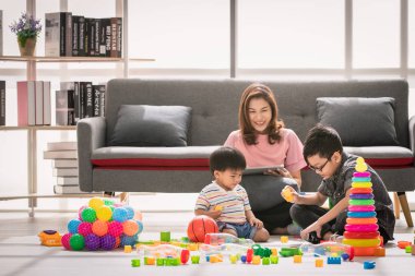 Asya Aile, boş zaman evde, genç güzel annem, rahatlatıcı, tablet kanepe, iki çocuk birlikte katta parlak ışık, rahat oturma odası çok renkli oyuncakları ile oynarken zevk kullanma.