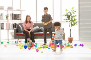 Asyalı aile, evde mutlu zaman, genç güzel anne, oğulları ile oynarken, küçük sevimli yürümeye başlayan, modern oturma odasında gözleri gözlük takan genç çocuk etrafında dağınık çok renkli oyuncaklar dolu