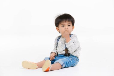 Stüdyo portresi, şirin, sevimli, Asyalı, kot pantolon giyen, uzun kollu tişört giyen, turuncu ayakkabılı, yerde bacakları açık oturan, izole edilmiş beyaz arka planda atıştırmalık yemekten hoşlanan çocuk.