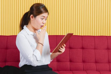Genç, çekici, Asyalı girişimci iş kadını portresi, modern ofisteki büyük kırmızı koltukta oturan, büyük gözlü, beklenmedik ve büyük bir sipariş alımında müşterilerden çıkan bir ifade.