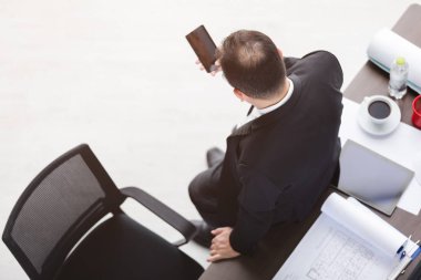 Üst görünüm, siyah takım elbiseli Asyalı iş adamı arka yüzü, masaya karşı duran, yoğun bir şekilde elinde akıllı telefon kullanmak, kırtasiye ile, beyaz güvenlik kaskı, dizüstü bilgisayar, tablet, akıllı telefon, ofis arka plan