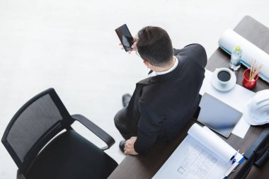 Üst görünüm, siyah takım elbiseli Asyalı iş adamı arka yüzü, masaya karşı duran, yoğun bir şekilde elinde akıllı telefon kullanmak, kırtasiye ile, beyaz güvenlik kaskı, dizüstü bilgisayar, tablet, akıllı telefon, ofis arka plan