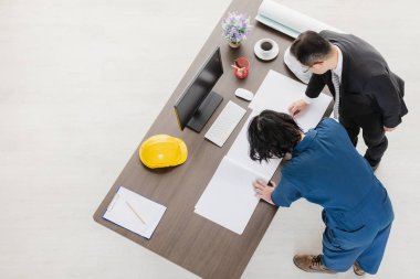 Asyalı iş adamı ve Asyalı erkek ustabaşının üst düzey görüşü, çalışma planı üzerinde ayakta ve eğilerek masaüstü, miğferler ve ofis geçmişi ile yeni proje hakkında tartışırlar.
