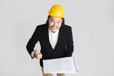 Mutlu, siyah takım elbise ve beyaz gömlek genç Asya iş adamı üst görünümü, sarı kask giyen, kameraya yukarı bakıyor, neşeyle thumb-up, elinde proje planı tutun, beyaz izole arka plan üzerinde