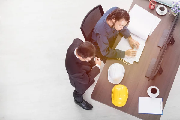 Top view of Asian business man point at desktop screen to Asian male foreman in coverall, sitting on chair, discuss about new project, with building plan and helmets and office background