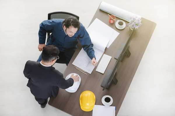 Top view of Asian business man point at desktop screen to Asian male foreman in coverall, sitting on chair, discuss about new project, with building plan and helmets and office background