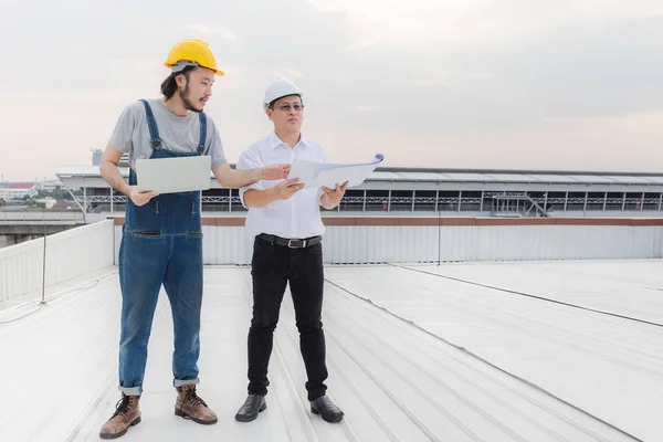 Middle age Asian factory manager and young Asian engineer in jeans jumpsuit, safety shoes, helmet, point something on far away on the rooftop of manufacturing plant
